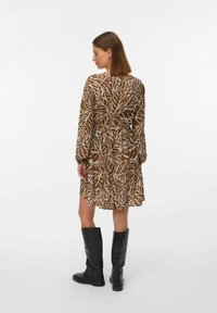 Robe marron avec motif de feuilles blanches, manches longues, taille ceinturée et ourlet évasé, associée à des bottes en cuir noir jusqu'au genou.