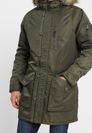 Mężczyzna ubrany w zieloną zimową parkę z kapturem wykończonym futrem, z wieloma kieszeniami na suwak i guziki oraz niebieskie dżinsy, ręka w kieszeni.