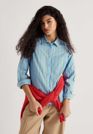 Frau mit langen lockigen Haaren trägt ein blau gestreiftes Hemd und beige Hosen, mit einem roten Strickpullover um die Taille gebunden, hält die Enden des Pullovers in der Hand.