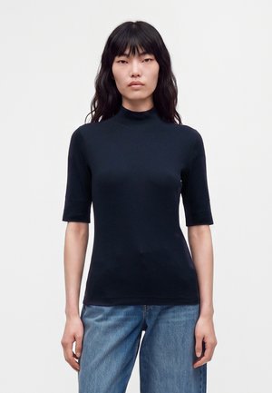 Femme aux longs cheveux noirs portant un col roulé noir ajusté à manches courtes et un jean bleu, debout devant un fond clair uni.