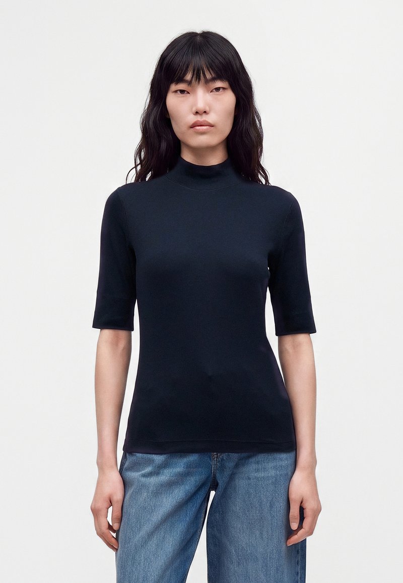 Femme aux longs cheveux noirs portant un col roulé noir ajusté à manches courtes et un jean bleu, debout devant un fond clair uni.