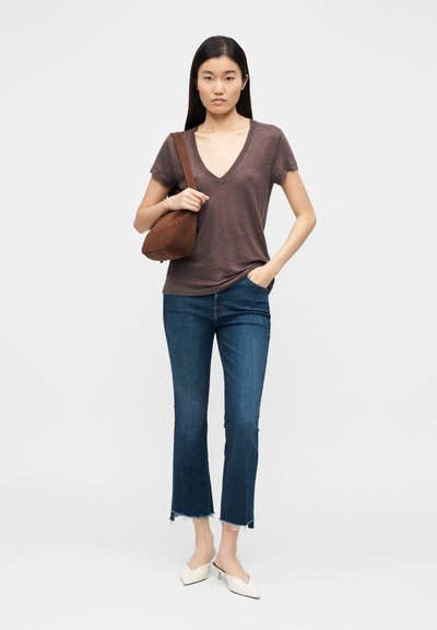 T-shirt castanha com decote em V, jeans azuis curtos com bainhas desfiadas e mules brancas; segurando uma mala castanha em camurça ao ombro. Fundo neutro.