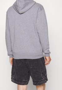 Persona che indossa una felpa grigia con cappuccio e pantaloni corti neri sbiaditi, vista di spalle su uno sfondo semplice.
