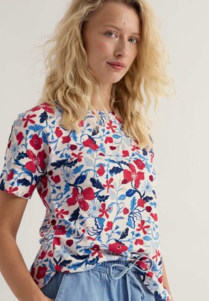 Donna bionda che indossa una maglietta bianca a maniche corte con stampa floreale rossa e blu, abbinata a pantaloni azzurri con coulisse.