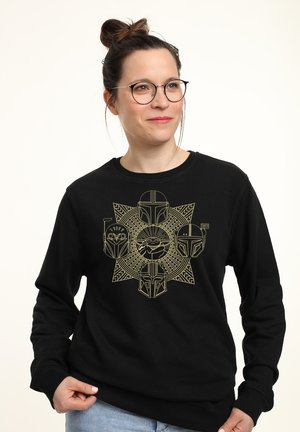 Kvinde med briller og knold i håret iført en sort sweatshirt med guld Star Wars Mandalorian hjelme og Baby Yoda motiver.