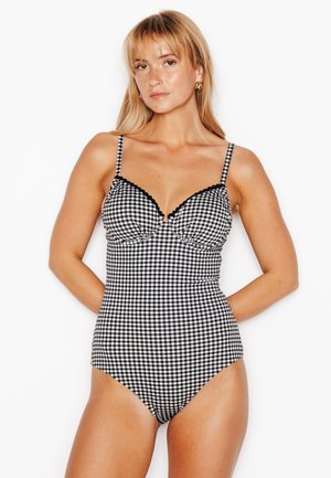 Femme aux cheveux blonds portant un maillot de bain une-pièce à carreaux noir et blanc avec des bretelles fines, debout sur un fond blanc.