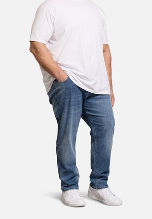 Homme portant un t-shirt blanc, un jean bleu, des baskets blanches, debout avec une main dans la poche devant un fond uni.