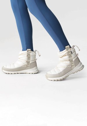 Hvide og beige isolerede støvler med polstret design, strukturerede såler og fremtrædende snøredetalje, båret med blå leggings.
