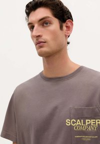 Grå bomulds t-shirt med rund halsudskæring, korte ærmer og brystlomme med gul trykdetalje. Enkel design, glat tekstur.