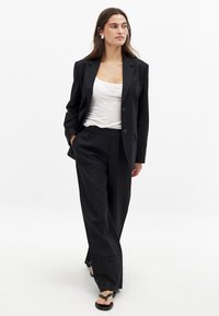 Čierny na mieru ušitý blejzer v kombinácii s voľnými čiernymi nohavicami s vyšívanými detailmi na leme. Modelka má na sebe biele tričko a čierne sandále.