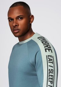 Sudadera azul claro con detalles en verde menta y texto negro en negrita "OMBRE EAT I SLEEP". Presenta un cuello redondo y mangas raglán.