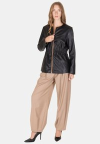 Giacca in ecopelle nera con vita arricciata, cerniera dorata, maniche lunghe, abbinata a pantaloni beige a gamba larga e tacchi neri.