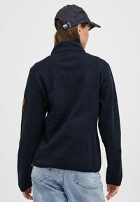 Marineblå fleecejakke med høy krage og lange ermer, med en brun logopatch på venstre arm og en glat tekstur.