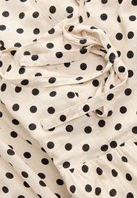 Abito di stoffa crema con pois neri, dotato di lacci e vita arricciata, realizzato in materiale leggero e di trama. 
