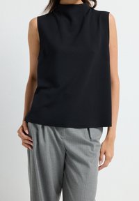 Femme portant un haut noir sans manches avec un col montant et un pantalon gris ample sur un fond uni.
