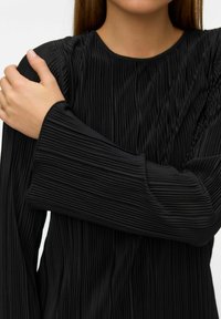 Czarna plisowana bluzka z długim rękawem i okrągłym dekoltem. Tkanina ma pionowe prążkowanie i luźny krój rękawów. Teksturowana powierzchnia.