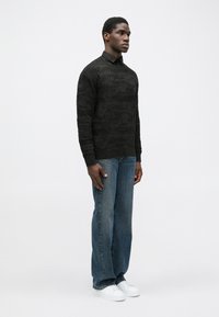 Pulover tricotat cu model negru, cu guler rotund, asortat cu blugi albaștri, largi, și adidași albi. Modelul stă de profil.