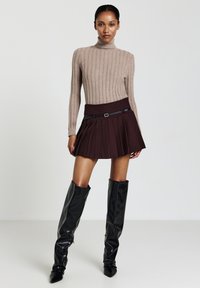 Femme portant un pull col roulé en tricot beige, une mini-jupe plissée noire avec une ceinture, et des bottes en cuir hautes noires sur un fond uni.