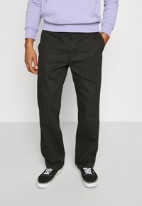 Man in zwarte casual broeken en zwarte sneakers met witte veters, hand in de zak, staande tegen een lichte effen achtergrond.