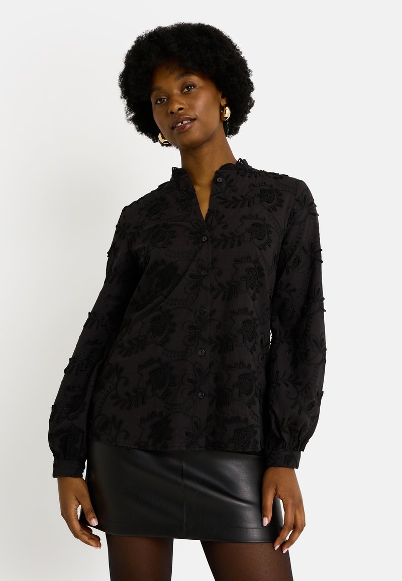 Blusa negra bordada con cuello mandarín, mangas largas con puños fruncidos y patrones florales en toda la prenda. Llevada con una falda de cuero negra.