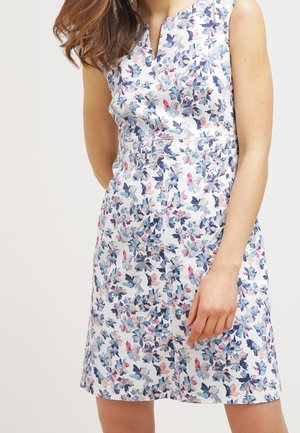 Robe blanche sans manches avec motif floral bleu et rose portée par une femme, longueur mi-cuisse et décolleté en V.