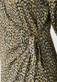 Tissu marron avec un motif de taches irrégulières en vert olive, blanc et noir. Comprend un détail de nouage latéral pour ajustement.