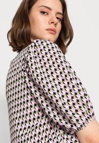 Top de mangas bufantes em tecido leve, apresentando um padrão geométrico em tons de rosa, verde e preto, com punhos elásticos franzidos.