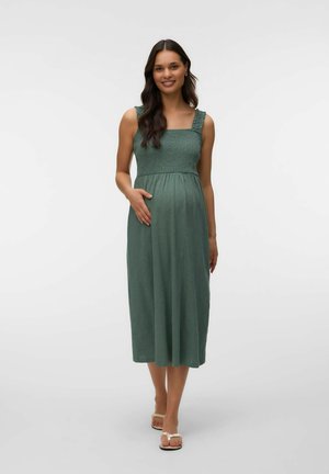 Pregnant woman with long dark hair wearing a sleeveless green midi dress and sandals, standing and smiling against a plain background.
