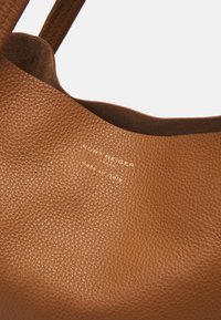 Hnědá kožená kabelka s texturovaným povrchem. Na boku je zlaté ražené logo "Kurt Geiger" a nápis "made in Italy".