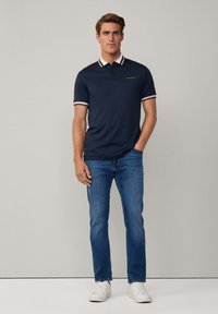 Námořnické polo triko s bílým a béžovým pruhovaným límcem, slim fit, s výšivkou loga. Spárováno s modrými džíny a bílými teniskami.