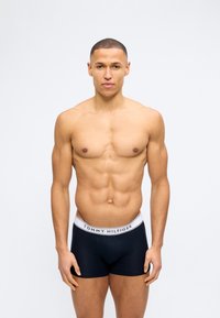 Μυώδης άνδρας στέκεται με το πρόσωπο προς τα εμπρός, φορώντας μαύρα μποξεράκια Tommy Hilfiger με λευκή μέση, σε απλό φόντο.