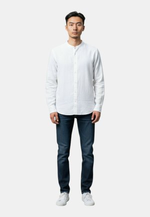 Uomo in piedi rivolto verso il fronte che indossa una camicia bianca abbottonata, jeans blu scuro e sneakers bianche su uno sfondo chiaro uniforme.