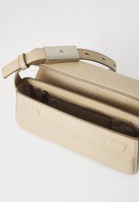 Beige lærveske med glatt tekstur, rektangulær form og metallisk detalj på stroppen. Inkluderer en glidelåsinnvendig lomme.