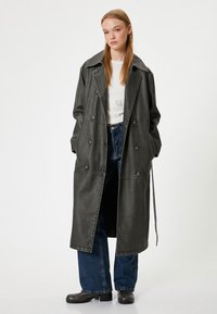 Long manteau en cuir noir avec un col classique, des boutons double boutonnage et des détails de couture visibles. Assorti à un jean bleu et des bottes noires.
