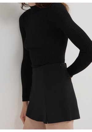 Femme portant un haut noir côtelé à manches longues et une jupe courte noire taille haute avec une fente devant.