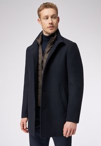 Cappotto di lana blu scuro con colletto alto, chiusura con bottoni e tasche laterali sopra un giubbotto imbottito marrone. Tessuto morbido e vestibilità sartoriale.