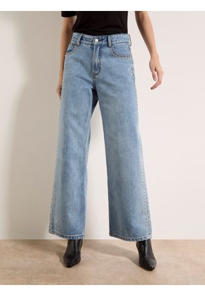 Donna che indossa jeans azzurri a gamba larga con borchie argentate lungo le tasche e le cuciture laterali, abbinati a stivali neri alla caviglia e top nero.