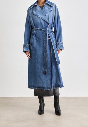 Femme portant un long trench en denim avec une ceinture nouée à la taille et des bottes à talons noires, debout sur un sol uni contre un mur blanc.