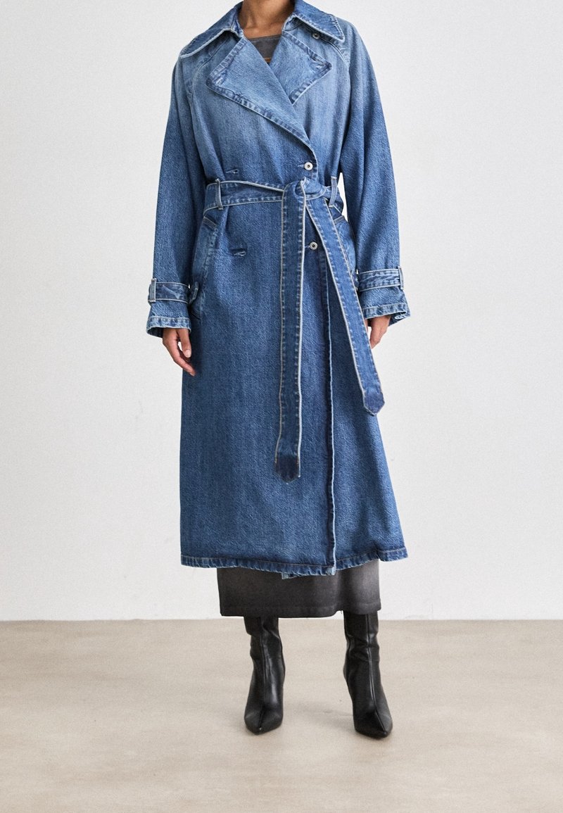 Femme portant un long trench en denim avec une ceinture nouée à la taille et des bottes à talons noires, debout sur un sol uni contre un mur blanc.