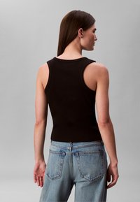 Vrouw met lang bruin haar, gekleed in een zwart mouwloos topje en lichtblauwe jeans met hoge taille, van achteren gezien tegen een grijze achtergrond.