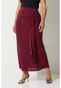 Burgunderröd maxikjol med vriden frontdetalj, gjord av slätt tyg. Kombinerad med svarta remklackar mot en enfärgad bakgrund.