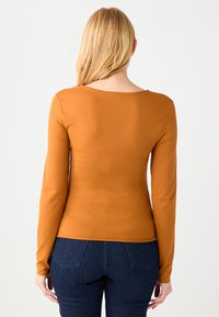 Koszula z długim rękawem w kolorze spalonym pomarańczu, dopasowany fason, gładka faktura, z okrągłym dekoltem i minimalnymi szwami, zestawiona z ciemnym dżinsem.