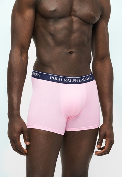 Μυώδης άνδρας φορώντας ανοιχτό ροζ boxer briefs Polo Ralph Lauren με μπλε ζώνη μέσης και λευκό λογότυπο, ποζάρει μπροστά σε απλό φωτεινό φόντο.