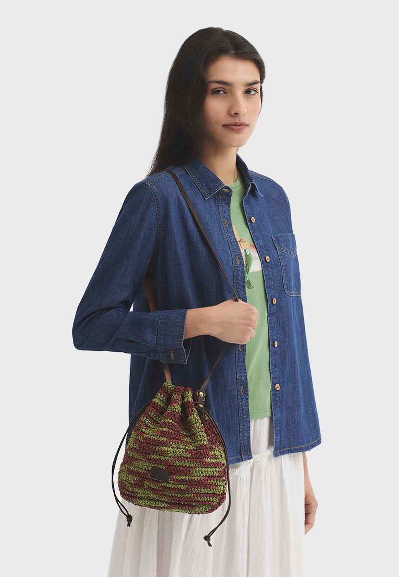 Donna che indossa una camicia di jeans blu sopra una t-shirt verde e una gonna bianca, con in mano una borsa a tracolla verde e marrone in maglia con coulisse.