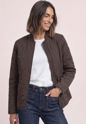 Vrouw die een bruine doorgestikte jas, wit t-shirt en blauwe spijkerbroek draagt, glimlacht en kijkt naar beneden met één hand in haar zak.