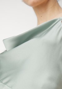 Grön satinblus med draperad halsringning, med mjuk, slät textur och subtil lyster. Nära vy som fokuserar på tygveck.
