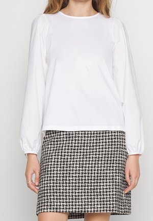Woman wearing a white long-sleeve blouse with gathered cuffs and a black-and-white houndstooth patterned skirt.