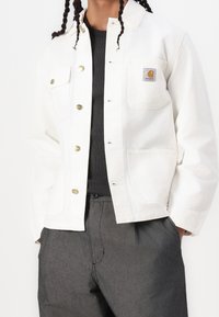 Chaqueta de lona blanca con botones de presión de metal, un bolsillo en el pecho con un parche de logo y un diseño relajado, combinada con una parte superior de canalé oscura.