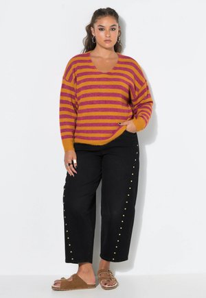 Frau mit langen Haaren trägt einen orange-lila gestreiften Pullover, schwarze Hosen mit goldenen Nieten und braune Sandalen und steht vor einer weißen Wand.