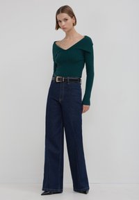 Top verde a costine con design off-shoulder, abbinato a jeans a gamba larga scuri in blu con vita alta e una cintura nera con hardware dorato.